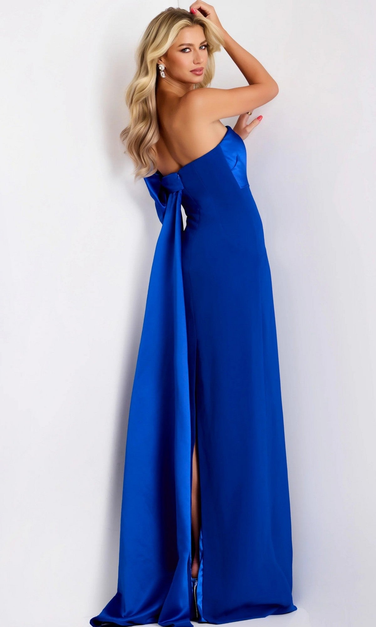 Woman in a bright blue strapless formal dress with a bow on the back stands against a white background, looking over her shoulder.